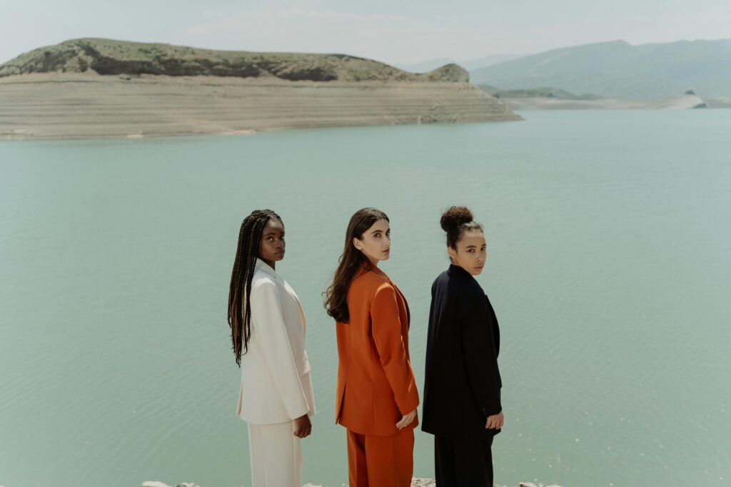 Three women in colorful blazers stand elegantly by a tranquil lake, exuding fashion and diversity.
